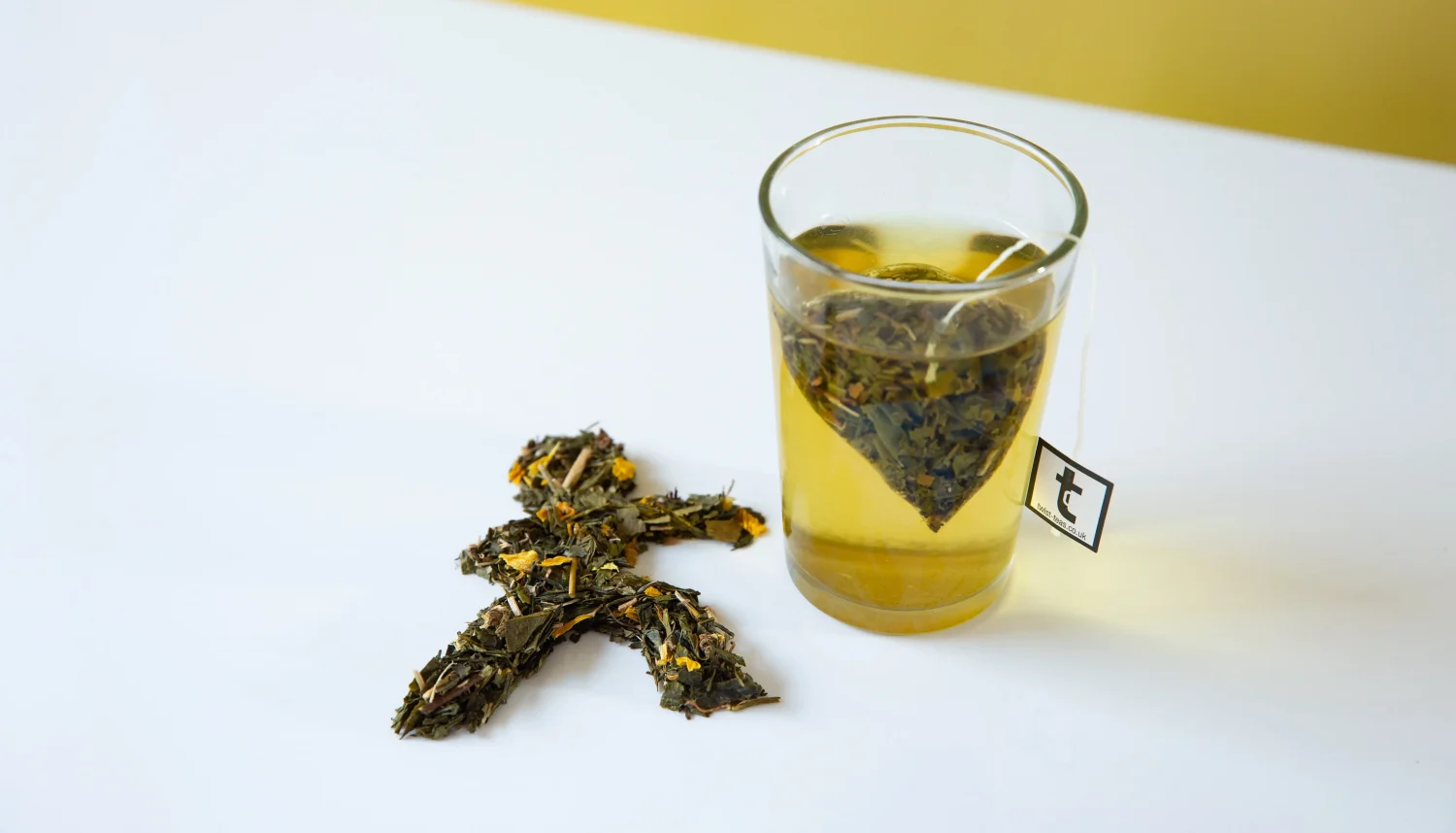 An image of a Green Teabag steeping in a glass. There are some Green Tea leaves personally arranged in the shape of a man next to the glass. 